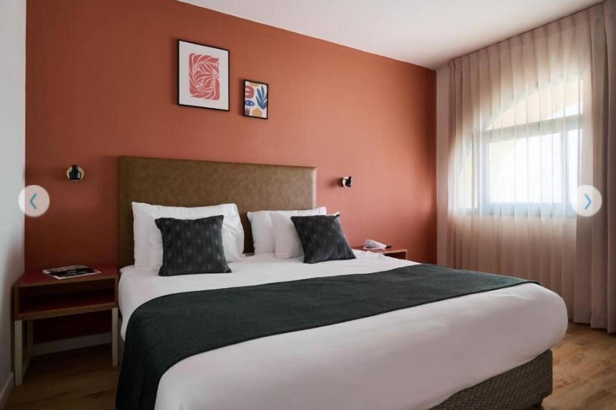 A hotel room with a large bed, a nightstand, a bedside table, a window with curtains, and two wall art pieces.