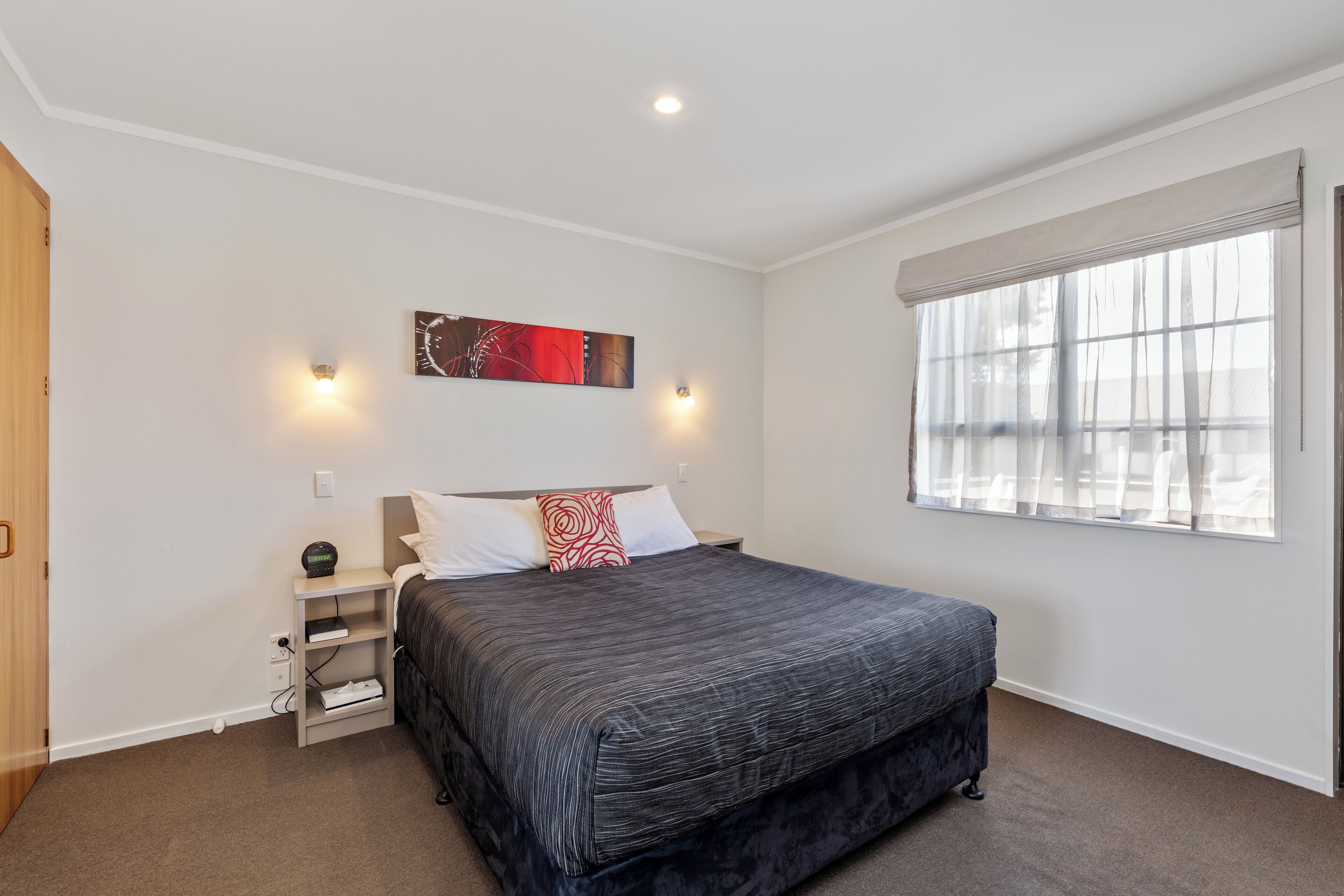 A bedroom with a large bed, a nightstand, a wall clock, and a window with sheer curtains.