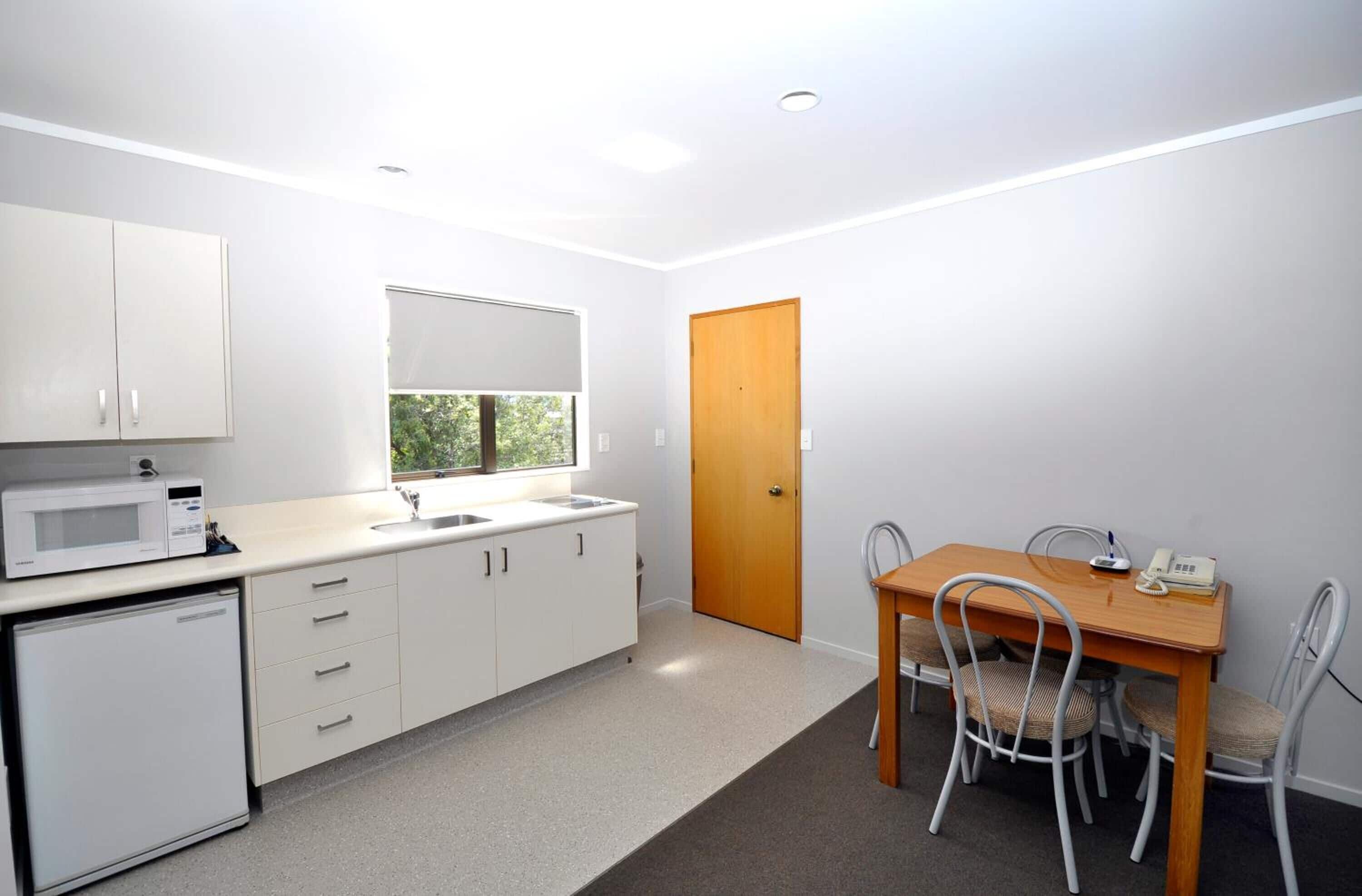 A compact kitchen with white cabinets, a microwave, a sink, and a small refrigerator. There's a wooden door and a dining table with chairs.
