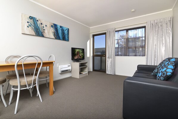 A modern living room with a wooden dining table, white chairs, a black leather sofa, and a television on a stand. - Knightsbridge Court Motor Lodge (Blenheim)