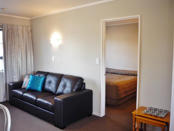 A living room with a black leather sofa, a blue pillow, and a wooden side table. - Knightsbridge Court Motor Lodge (Blenheim)