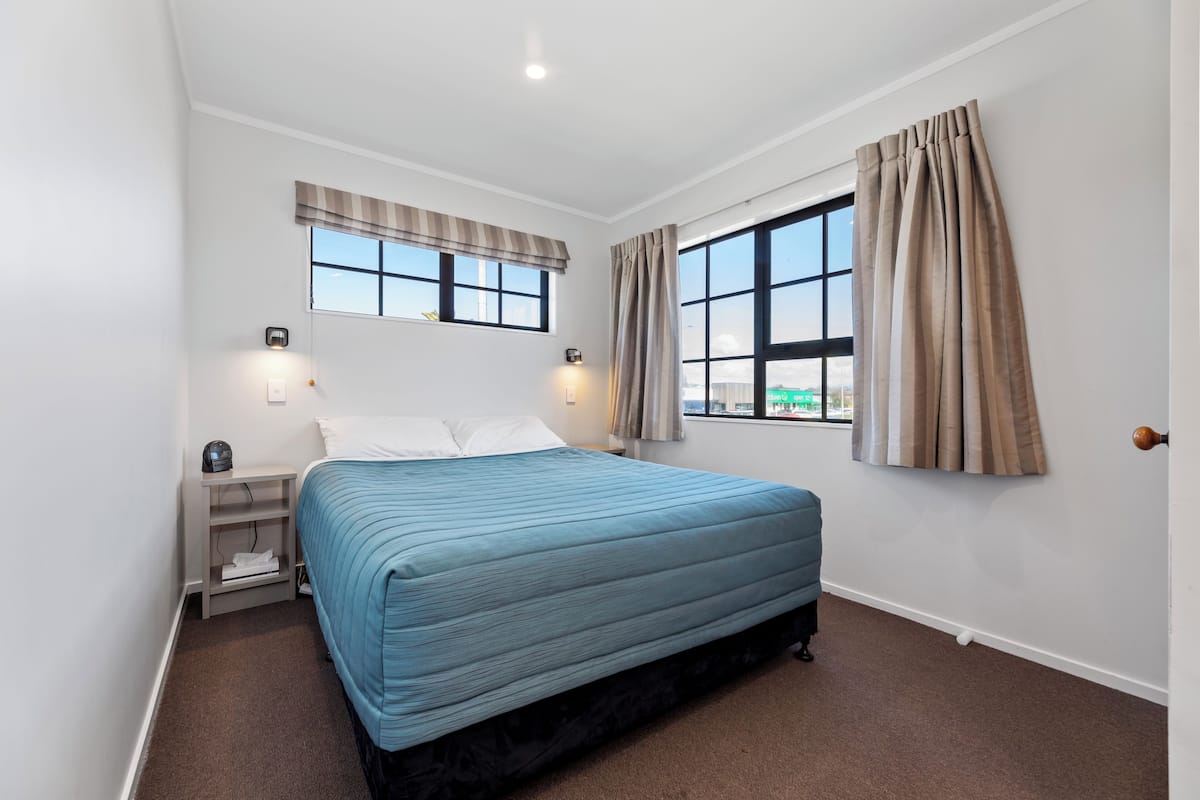 A bedroom with a bed, a nightstand, a window with curtains, and a view of the city.