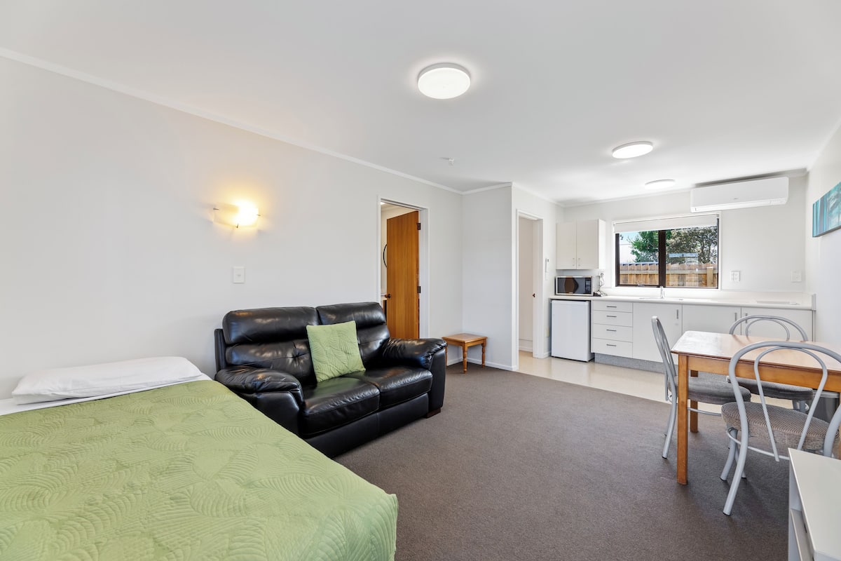 A modern, well-lit room with a green bed, a black leather sofa, a dining table with chairs, and a kitchen area.