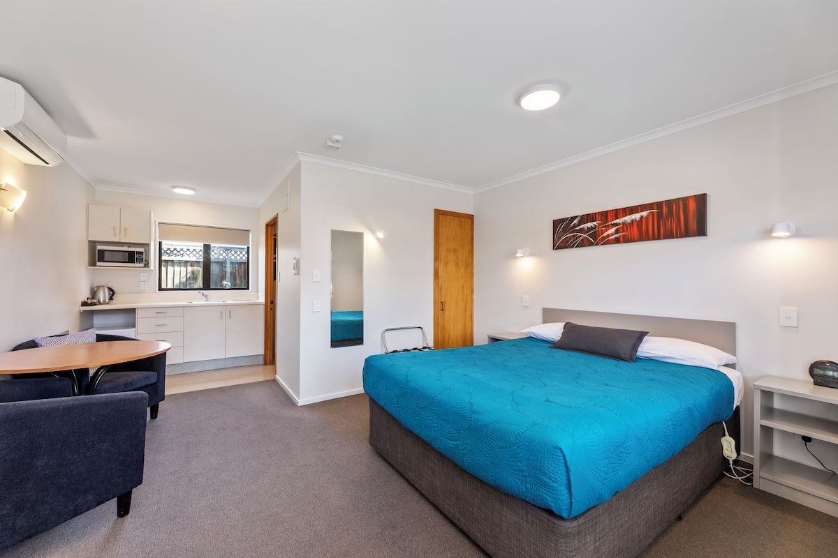 A modern hotel room with a large bed, a desk, and a kitchenette.