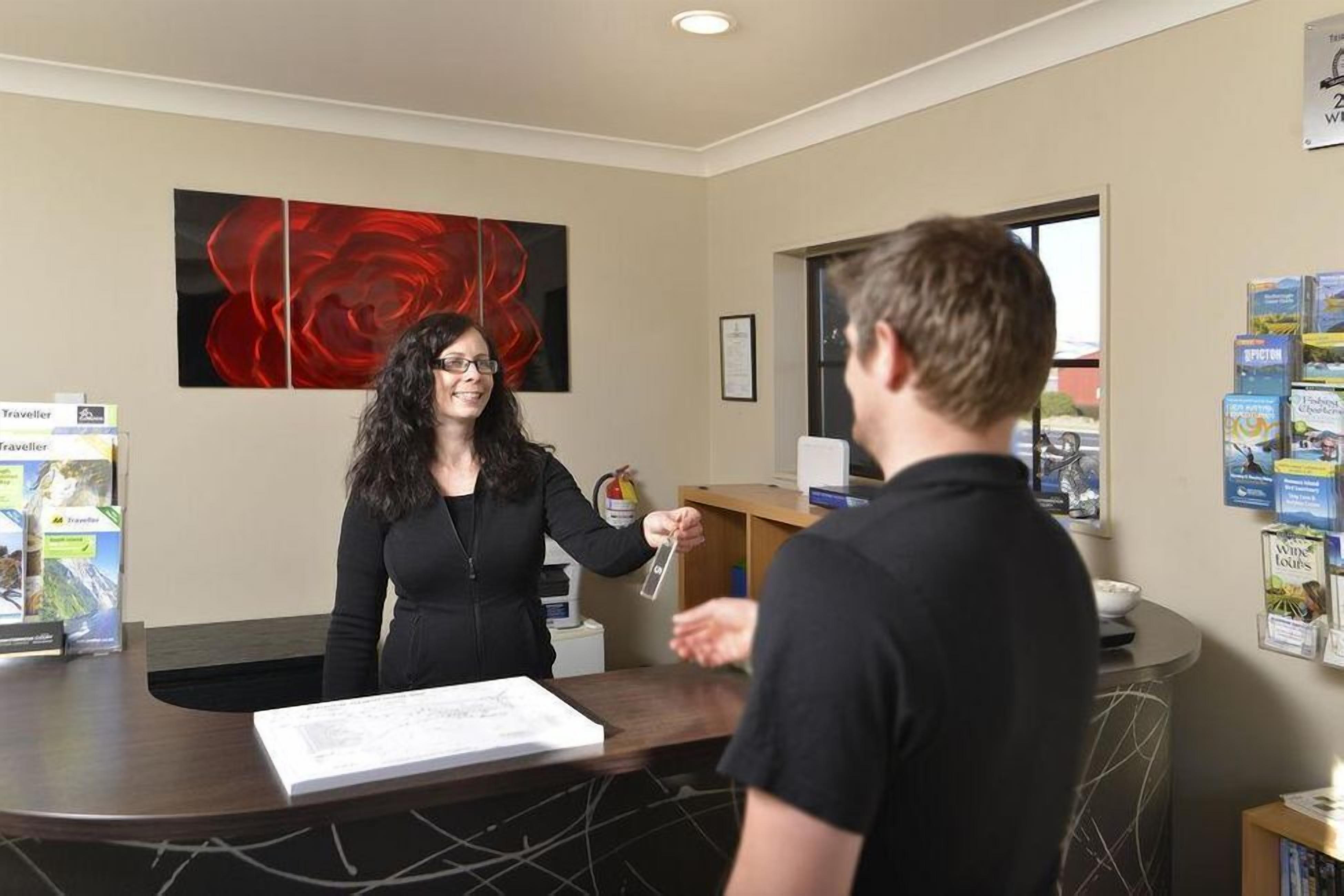 Een receptiebalie met een vrouw die een kaart vasthoudt, een man die voor haar staat en reisbrochures aan de muur.
