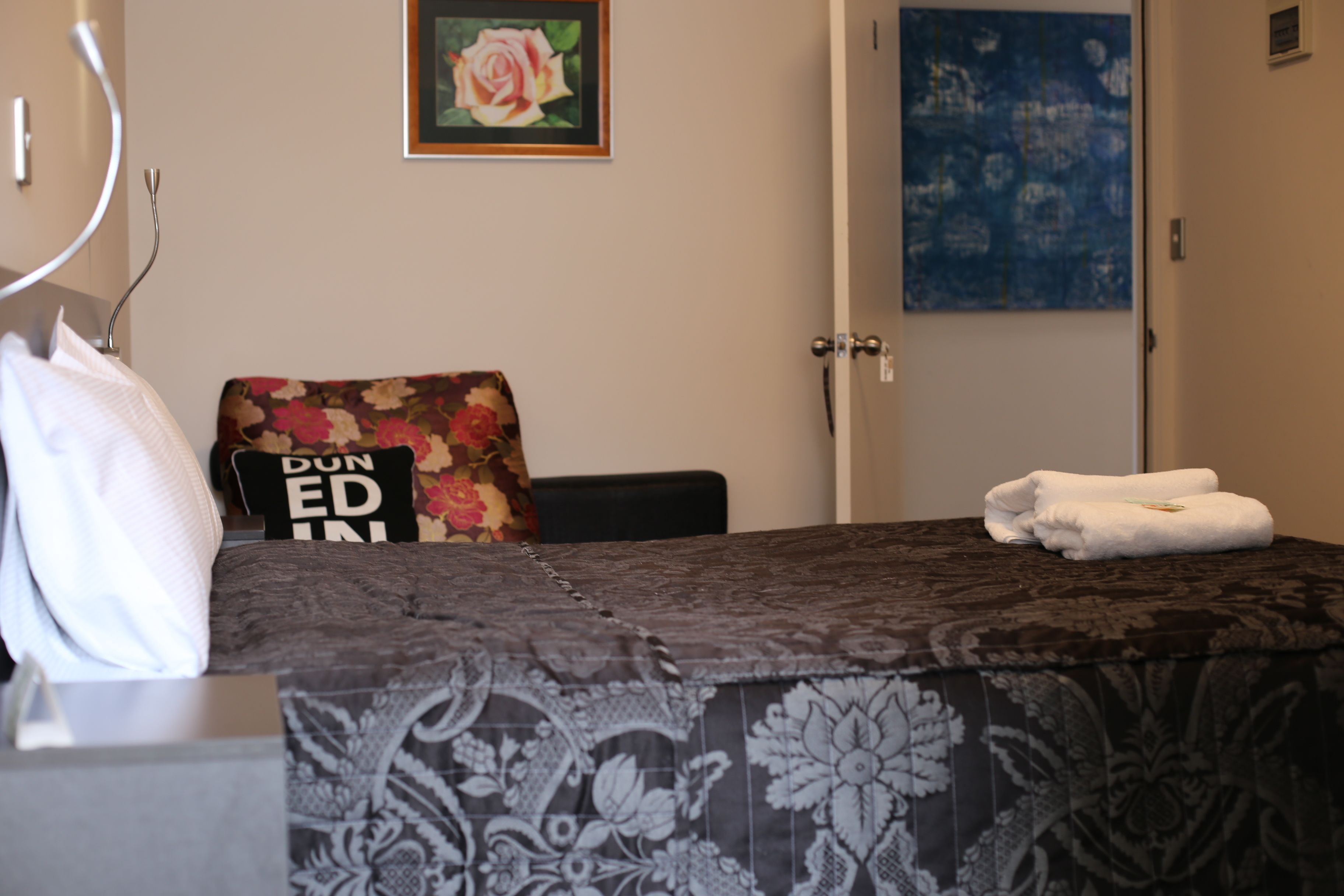 Quarto de hotel com uma cama bem arrumada, uma cadeira com almofada florida, um quadro com uma rosa e uma pintura abstrata azul na parede.
