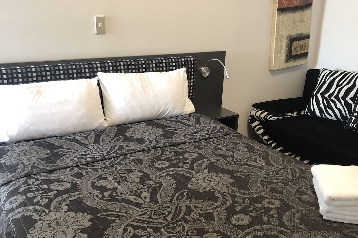A neatly made bed with a patterned bedspread, a bedside table with a lamp, and a black sofa with a patterned pillow.