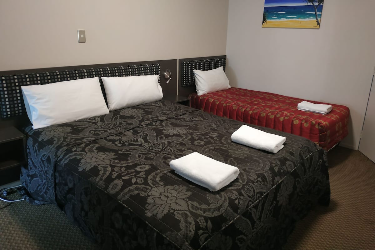 A hotel room with a large bed, two pillows, a red and black bedspread, a white pillow, a red and gold bedspread, and a painting of a beach on the wall.