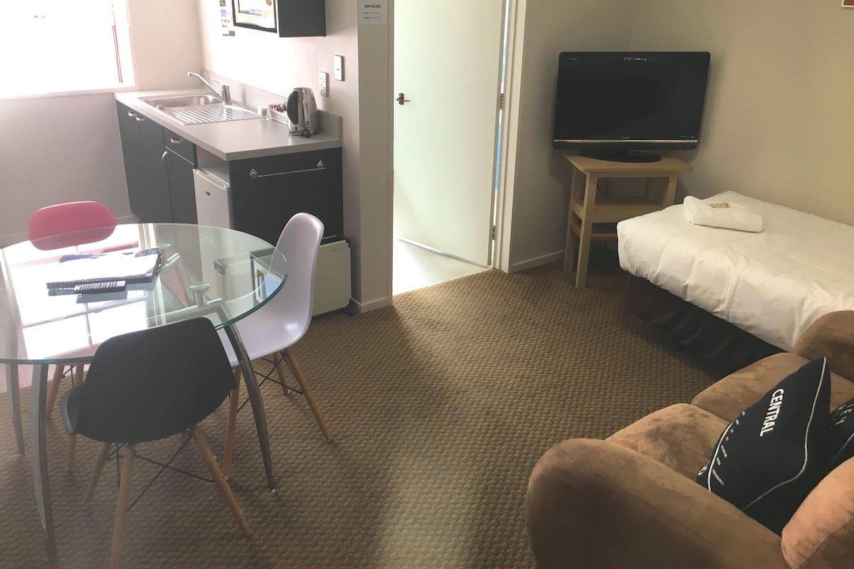 A compact hotel room with a kitchenette, a glass dining table with chairs, a single bed with a pillow, a television on a stand, and a carpeted floor.
