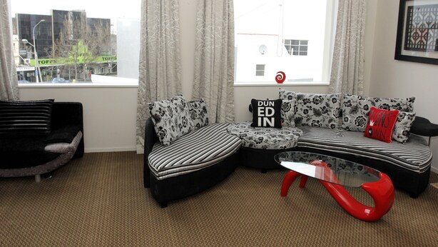 Un salon moderne avec un canapé d’angle, un pouf à motifs noir et blanc et une table basse en verre rouge.