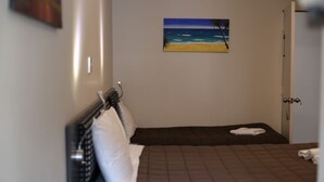 Un lit simple avec une couverture marron, un oreiller blanc et un tableau représentant une scène de plage accroché au mur.