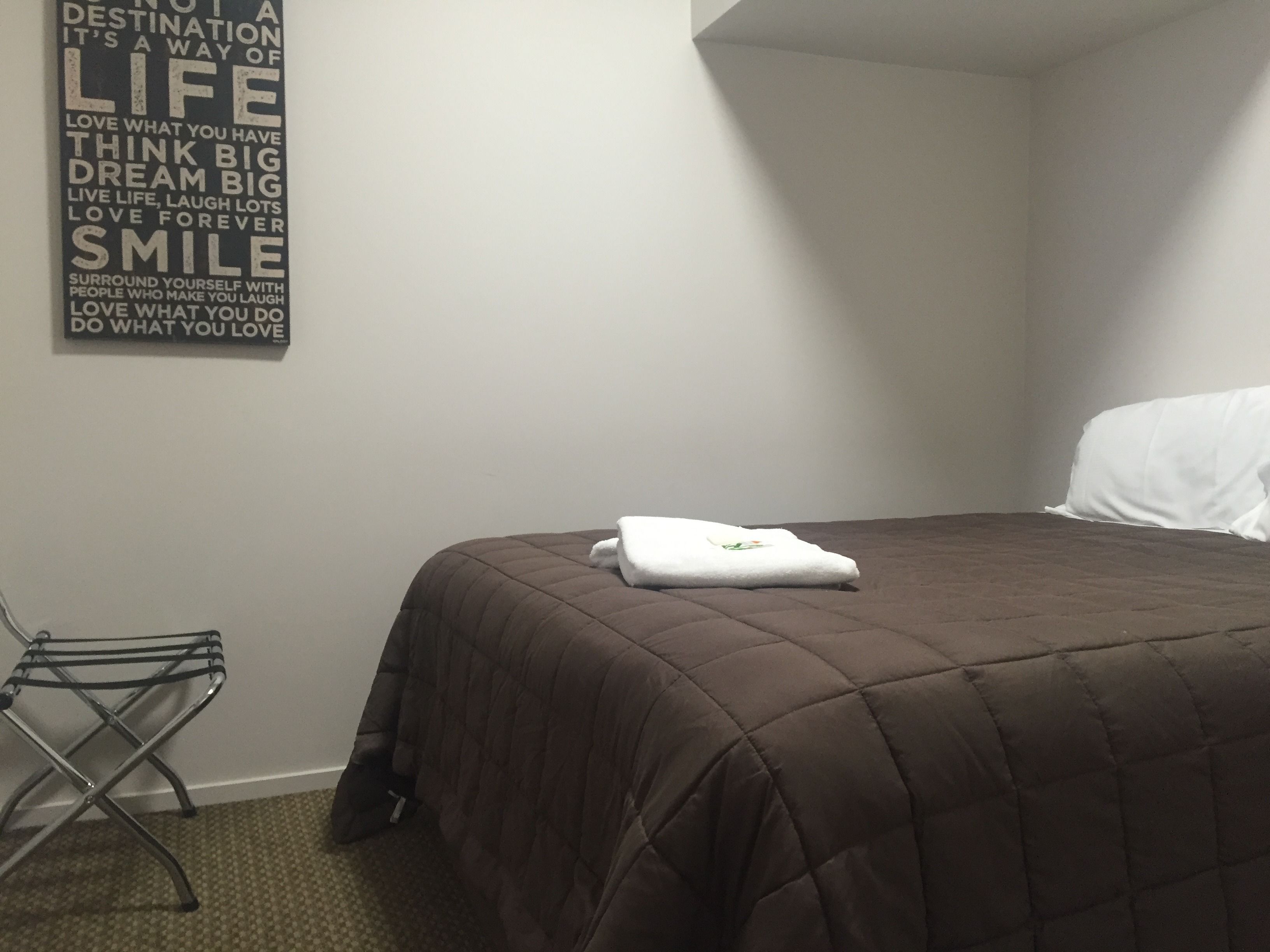 A neatly made bed with a brown quilt, a white pillow, and a folded white towel on the bed. There is a metal chair with a towel rack next to the bed. A motivational quote is displayed on the wall above the bed.
