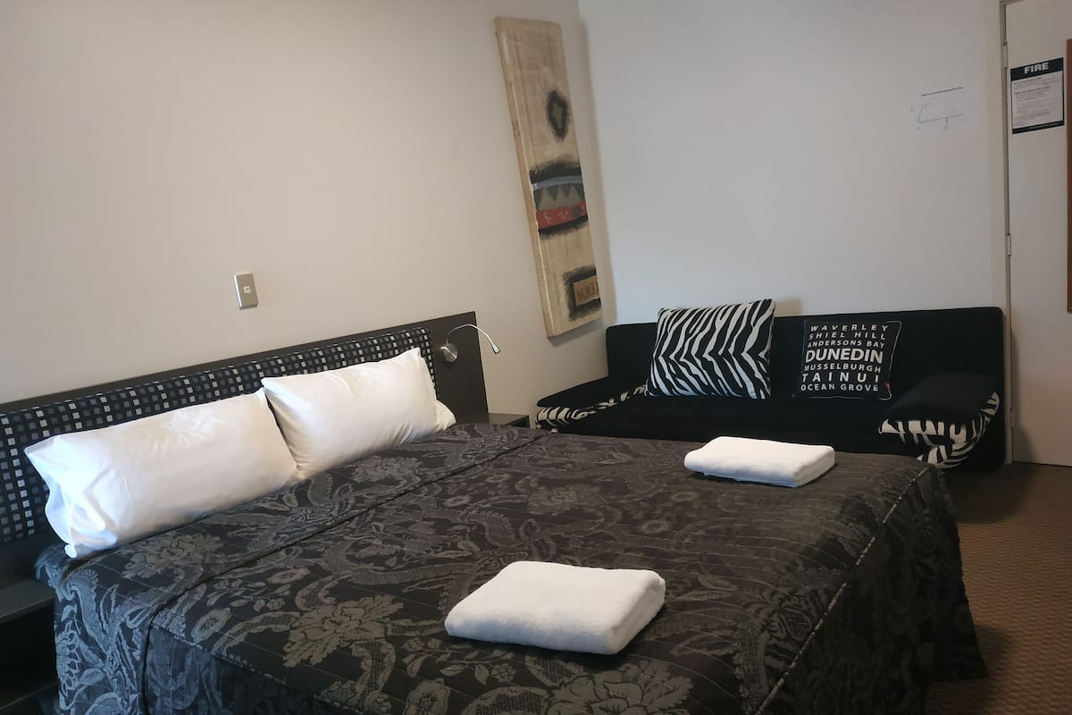 A hotel room with a large bed, two pillows, a black sofa, and a wall-mounted artwork.
