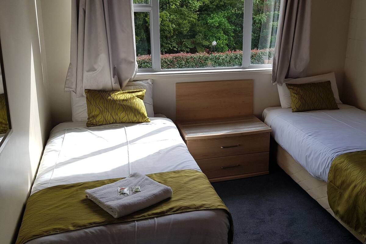A small, well-lit room with two single beds, a wooden nightstand, and a window with curtains.