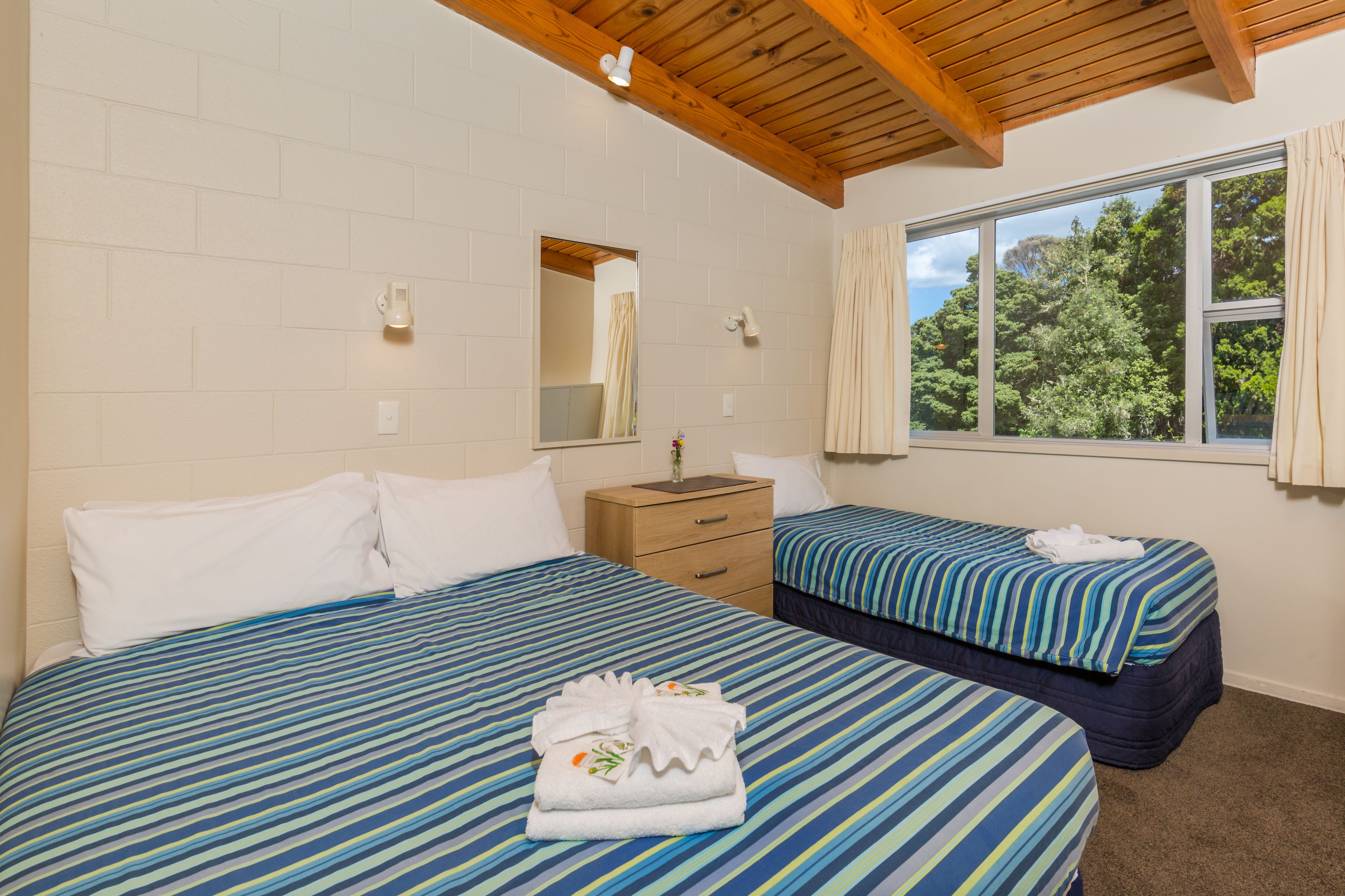A hotel room with two beds, a wooden ceiling, a window with curtains, a mirror, and a bedside table.