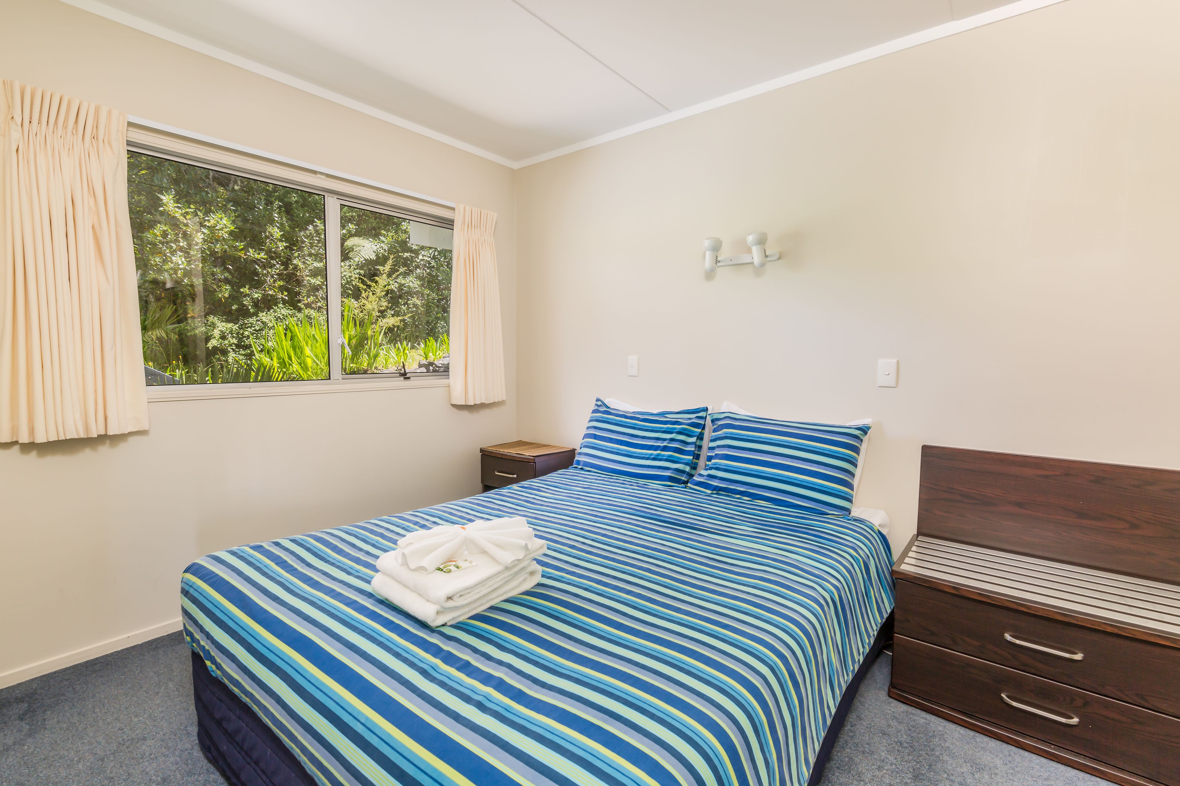 Ein Schlafzimmer mit Bett, Nachttisch, Schminktisch, einem Fenster mit Vorhängen und einer Wandleuchte.