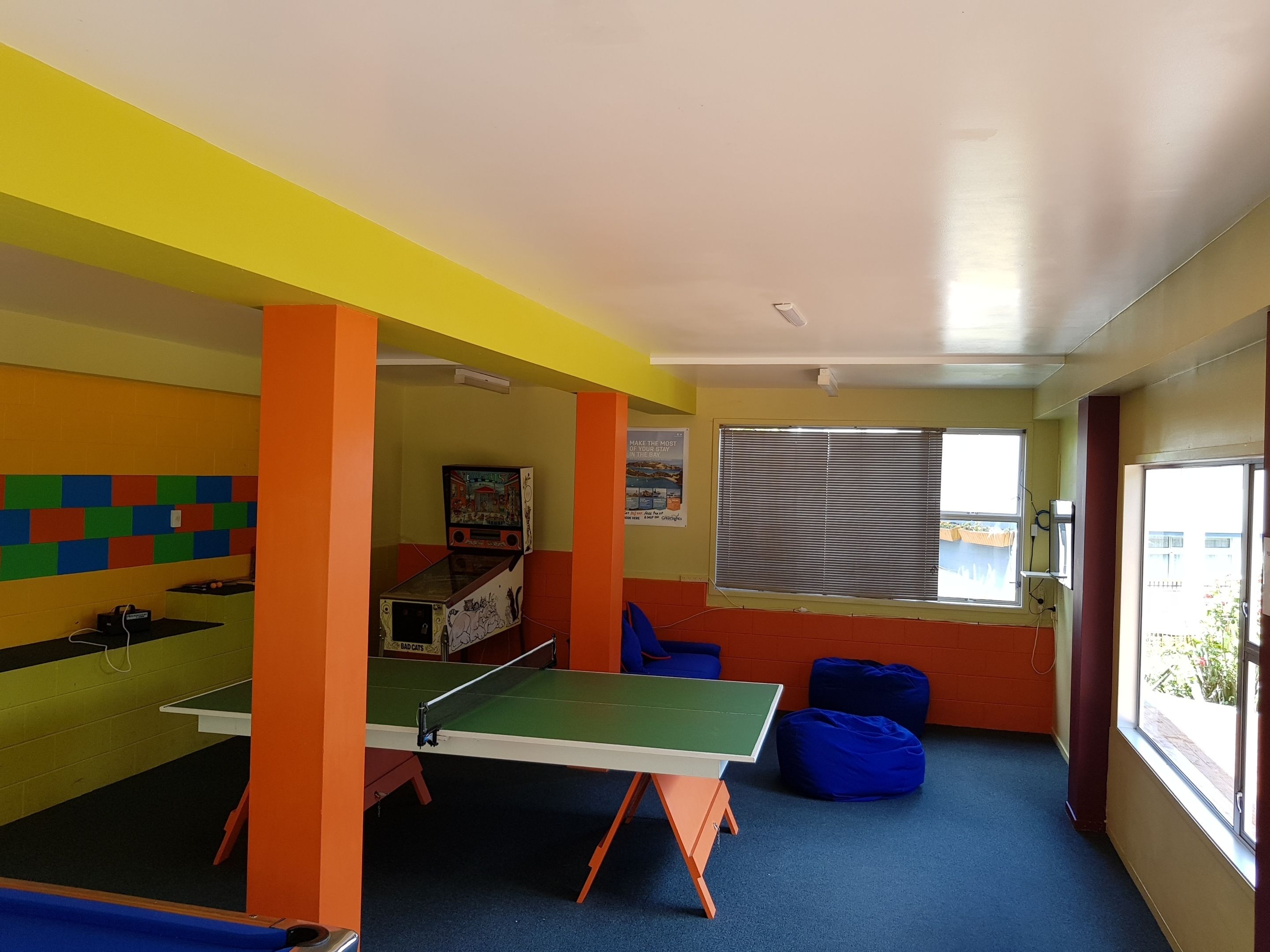 A room with a ping pong table, a small table with a video game, colorful wall tiles, a window with blinds, and blue bean bags.