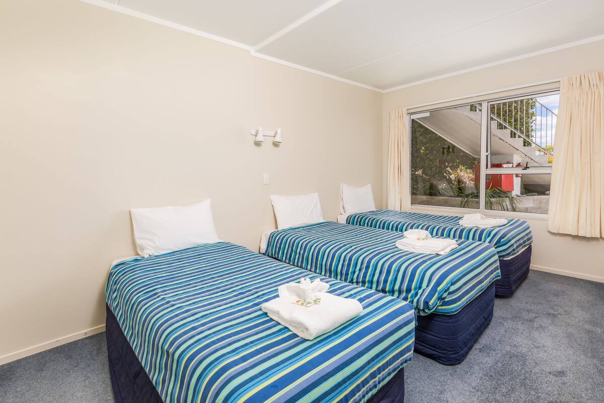 A hotel room with two beds, a large window with curtains, and a view of a staircase and outdoor area.