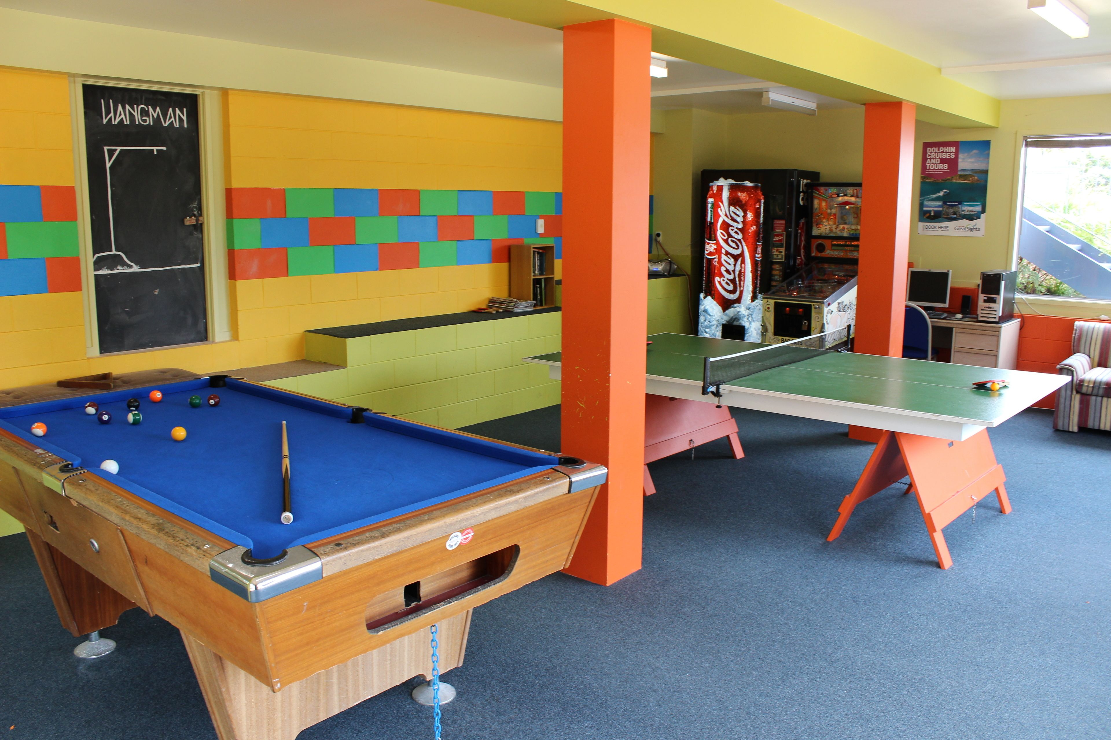 A pool table with balls, a dartboard, and a pinball machine in a room with colorful walls and orange pillars.