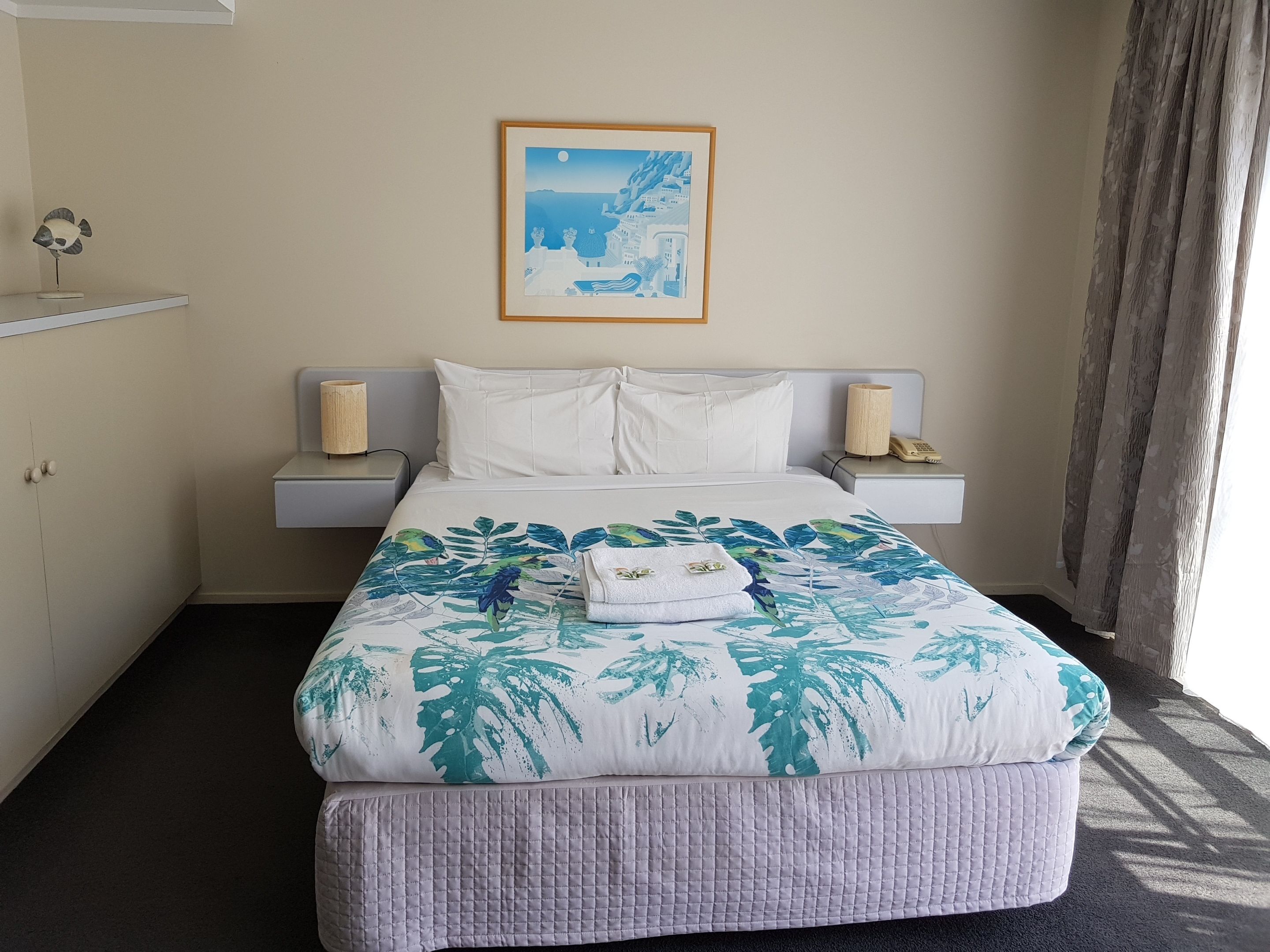 A hotel room with a bed, a nightstand, a lamp, a framed picture on the wall, and a window with curtains.