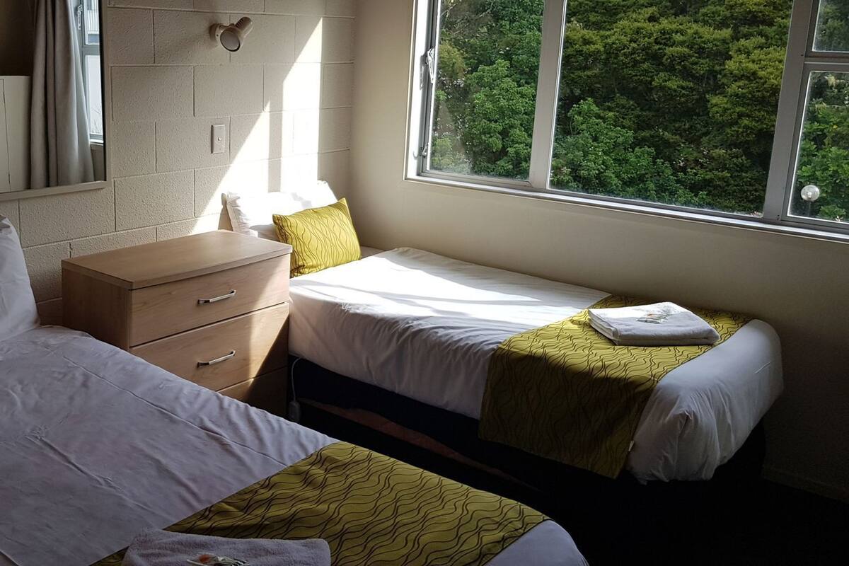 A hotel room with two beds, a wooden nightstand, a mirror, and a window with a view of trees.