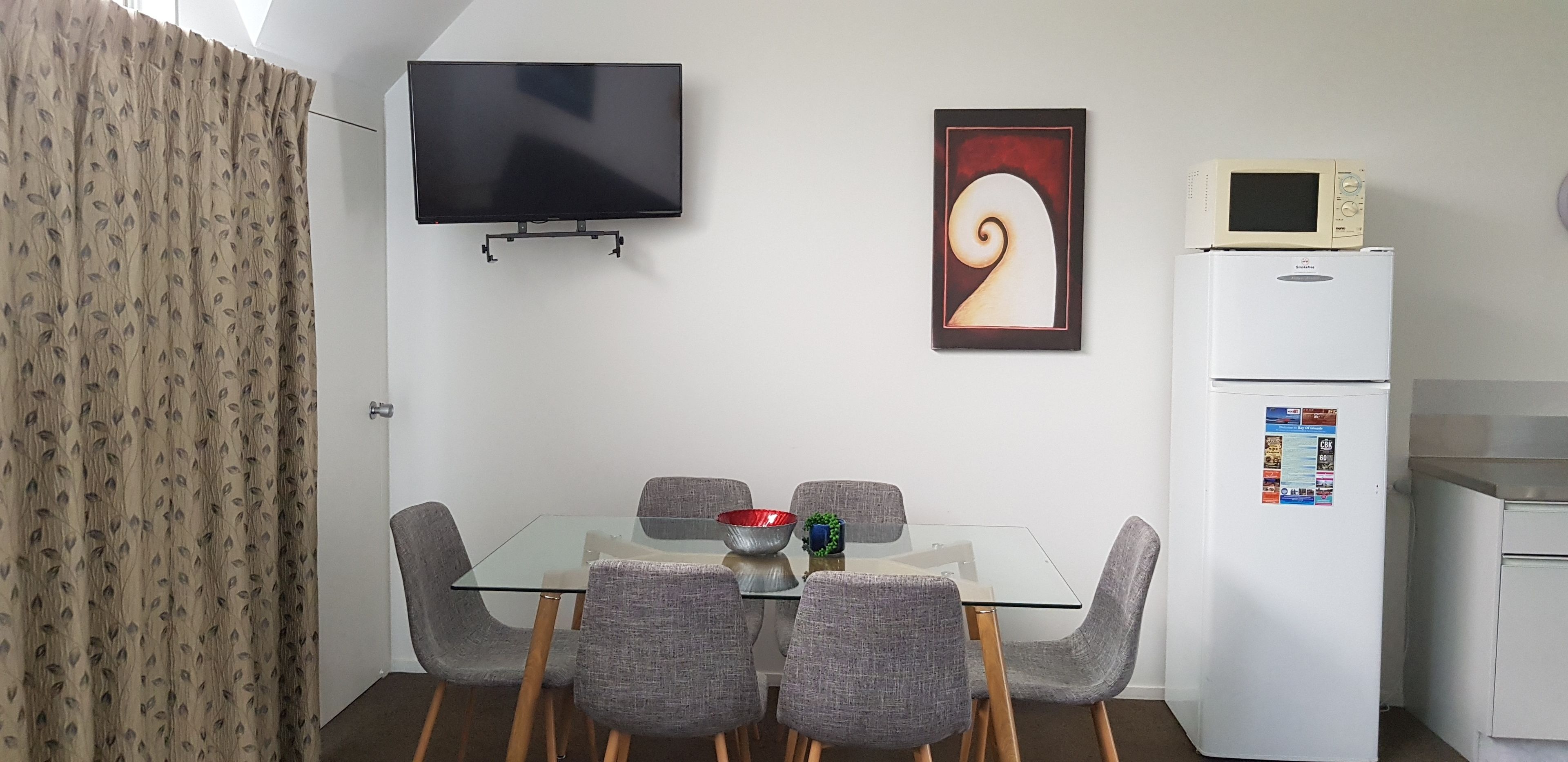 A compact room with a glass table, four grey chairs, a flat-screen TV, a microwave, and a refrigerator.