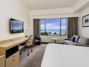 A hotel room with a bed, a desk, a chair, a sofa, a small table, and a large window with a view of the water.