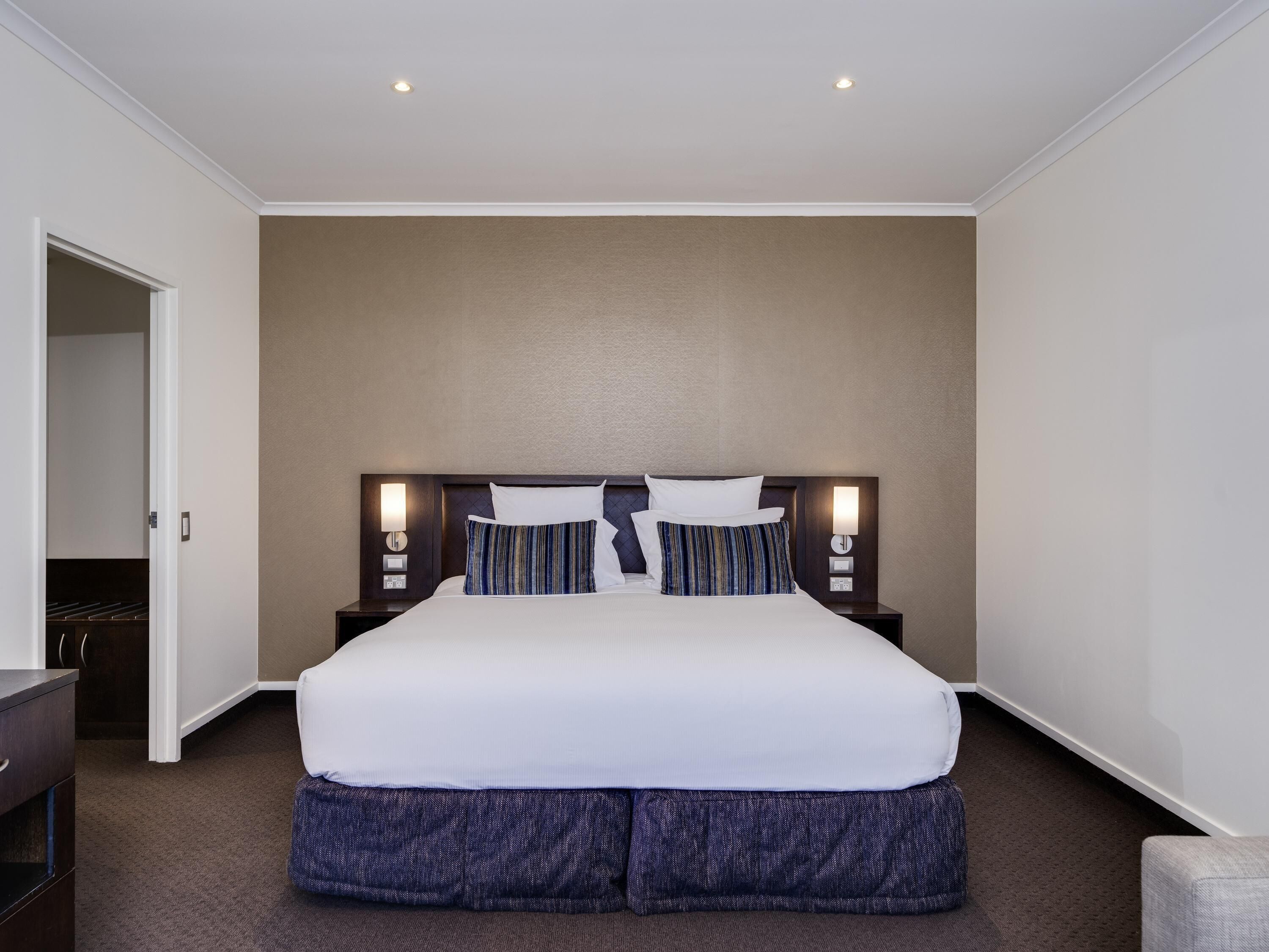 A hotel room with a large bed, two bedside tables with lamps, a wardrobe, and a door leading to another room.
