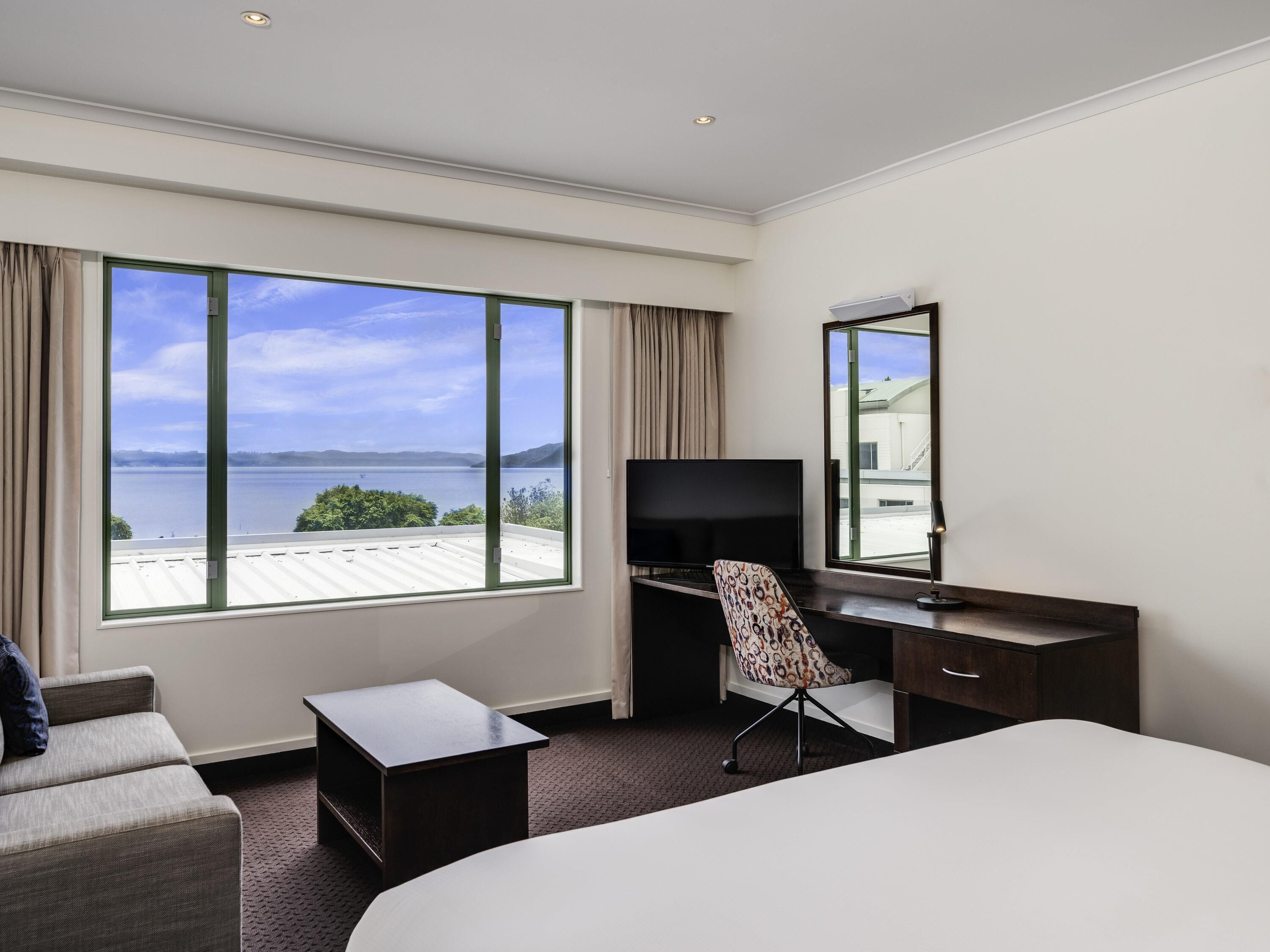 A hotel room with a large window offering a view of the sea and hills, a bed, a desk with a chair, and a television.