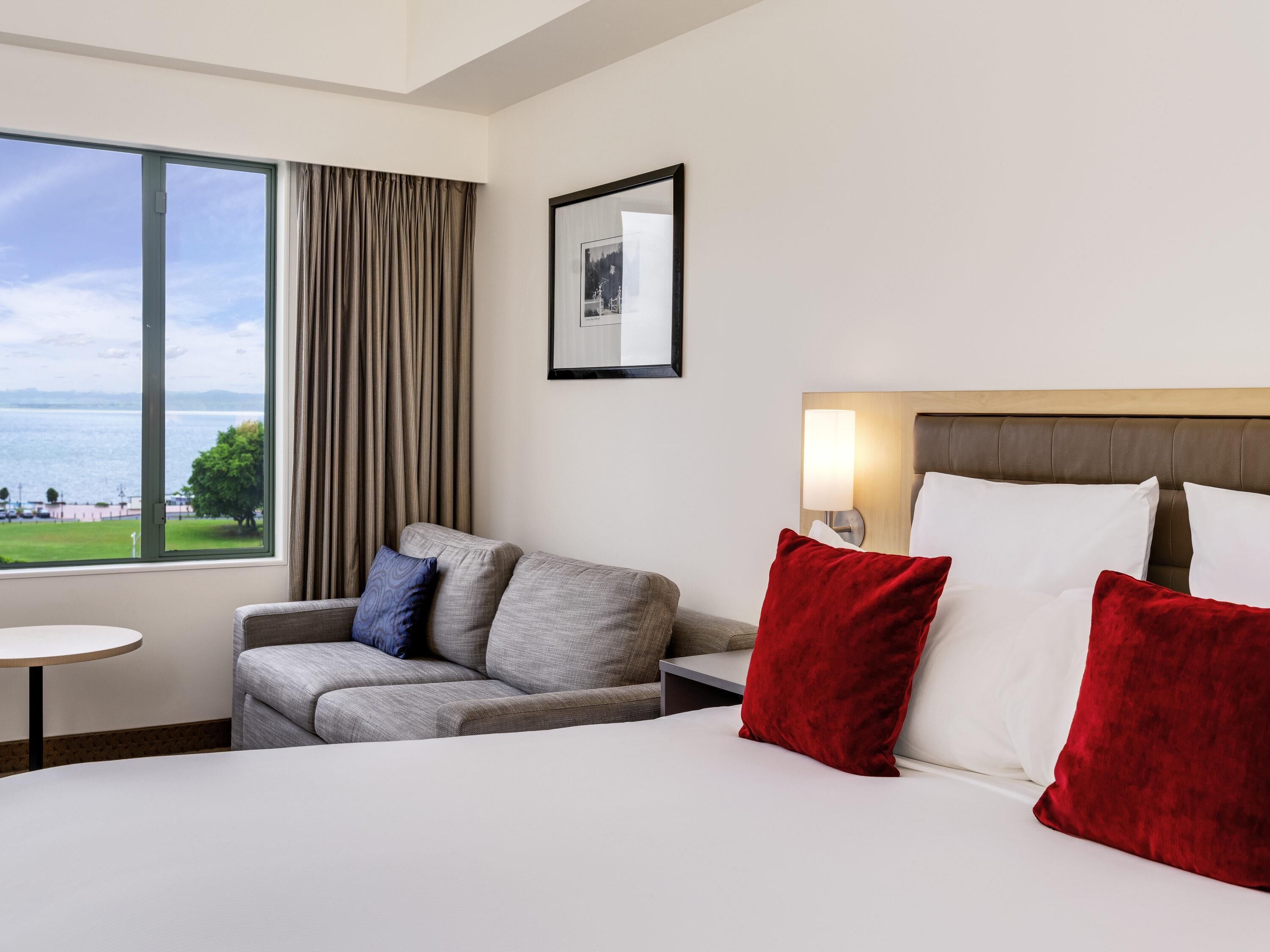 A hotel room with a bed, a sofa, a small table, a lamp, and a window with a view of the sea.