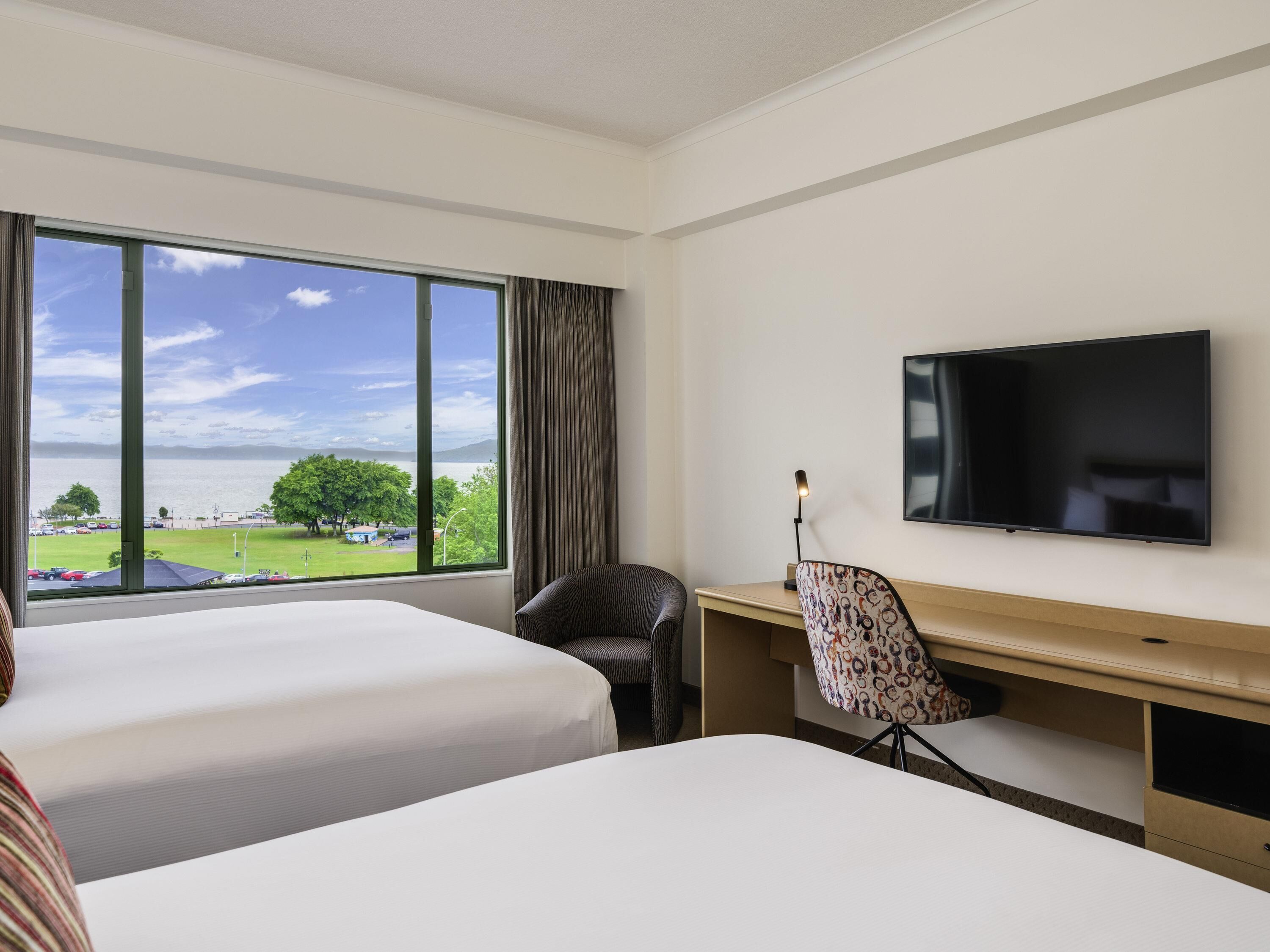 A hotel room with two beds, a desk, a chair, a TV, and a large window with a view of a park and water.