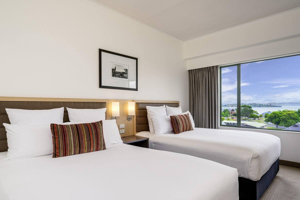 A hotel room with two beds, a large window with a view, a framed picture on the wall, and a nightstand with a lamp.