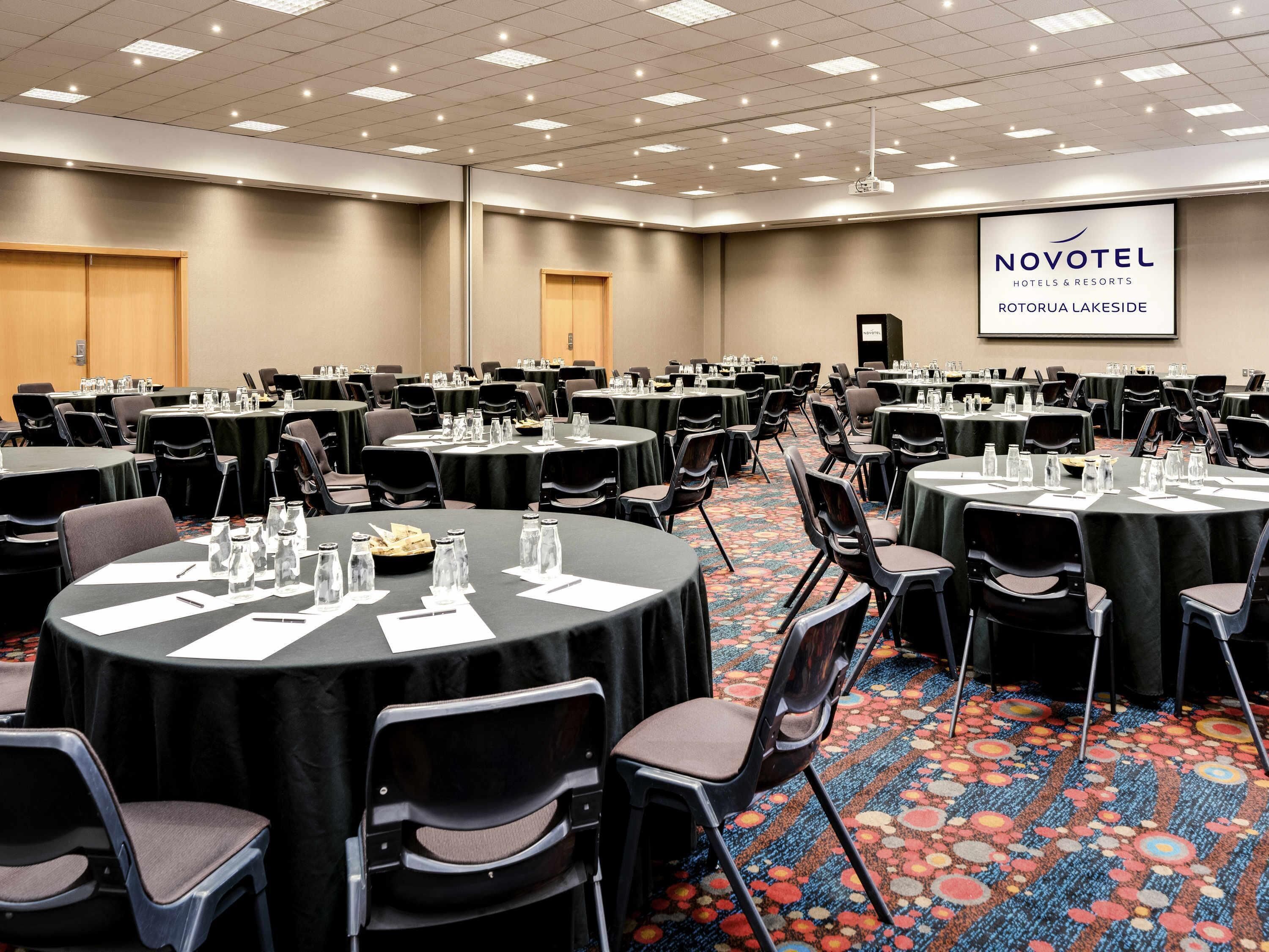 Une salle de conférence équipée de tables rondes, de chaises et d’un écran de projection affichant le texte « NOVOTEL HOTELS & RESORTS ROTORUA LAKESIDE ».