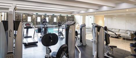 A gym with various exercise machines, a bench, and a pool area visible through the glass wall.