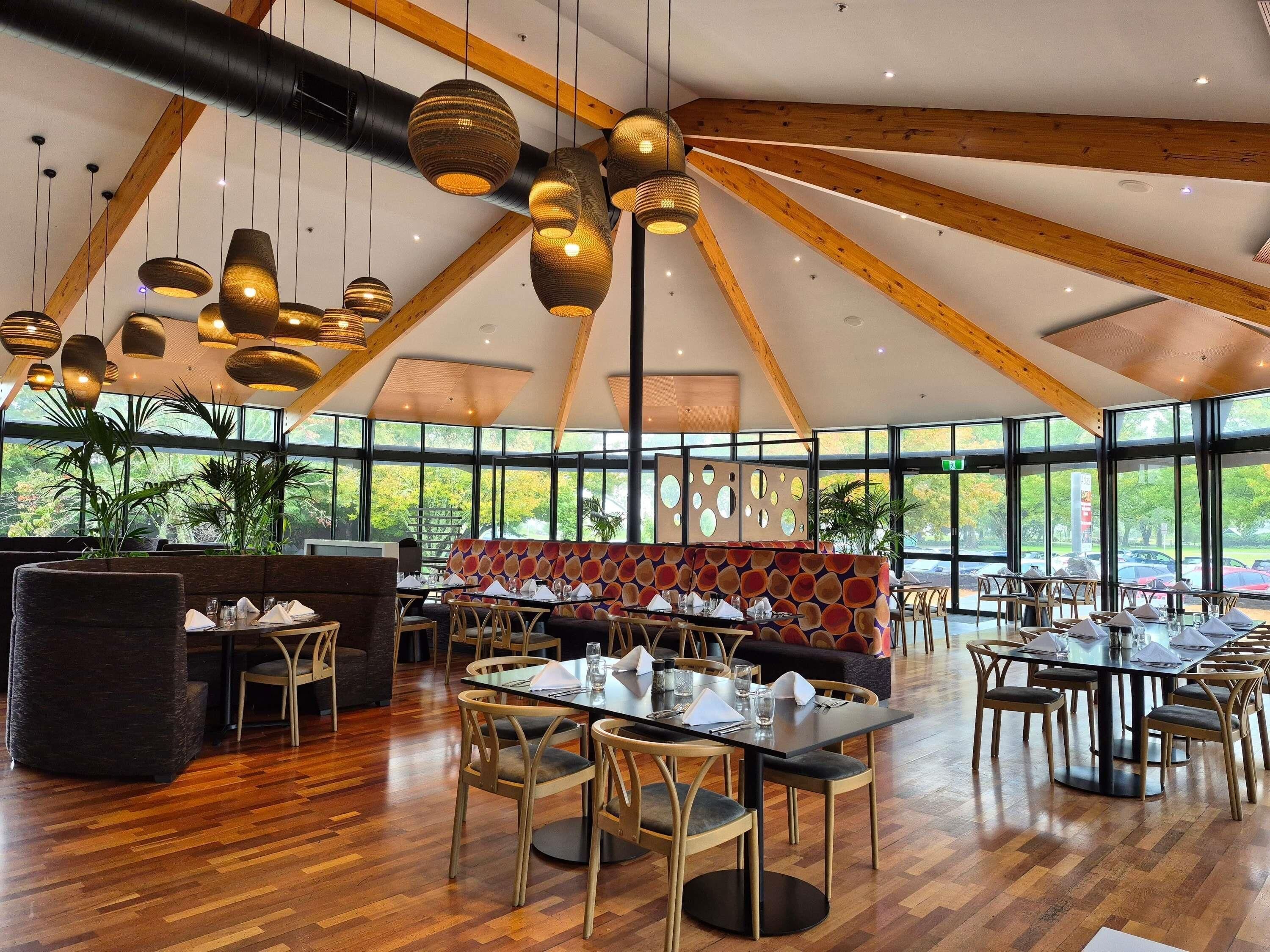 A spacious restaurant with high ceilings, wooden beams, and large windows offering a view of the outdoors. Tables are set with chairs, and there are hanging pendant lights.