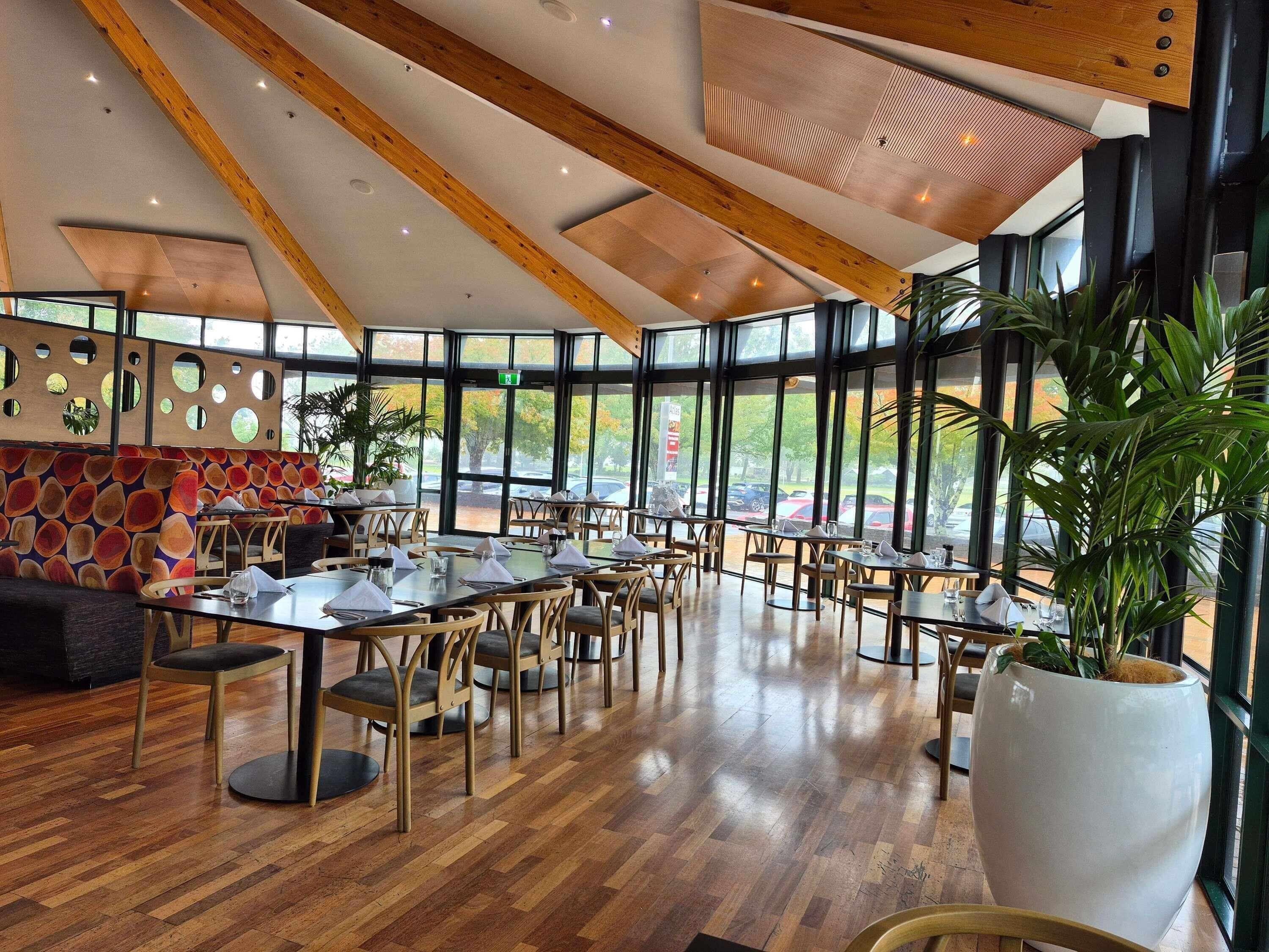 Un restaurante espacioso con techos altos, vigas de madera y grandes ventanales que ofrecen vistas al exterior. Las mesas están ordenadas con sillas, y hay plantas en macetas que añaden verdor.
