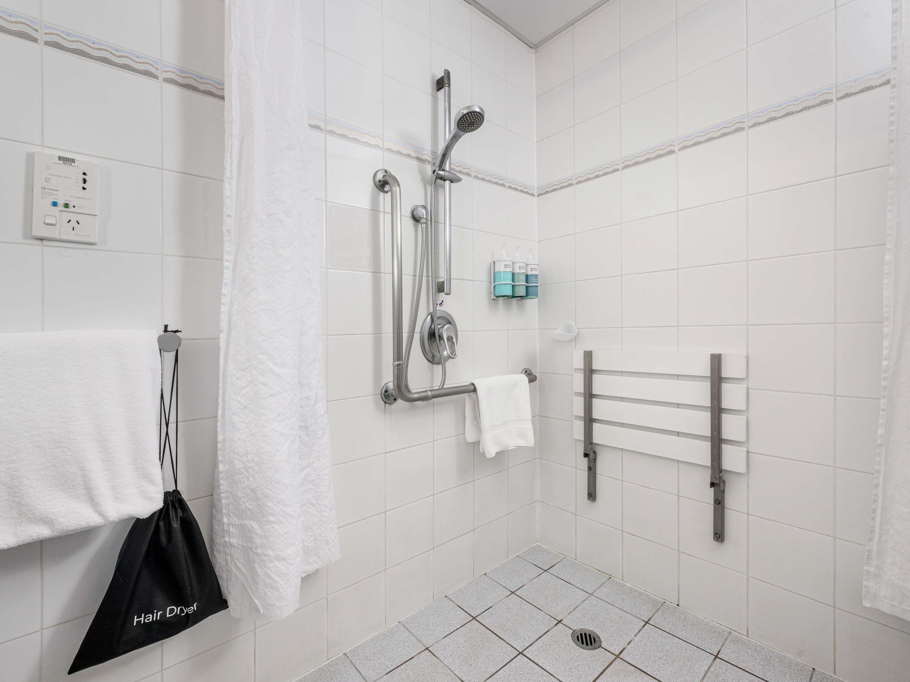 A modern bathroom with white tiles, a shower with a handheld showerhead, a towel rack, and a hair dryer bag.