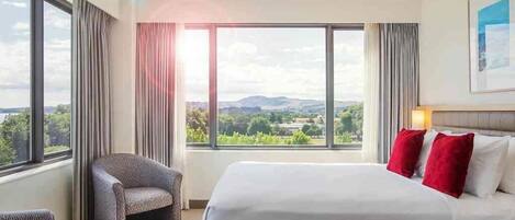 A hotel room with a large bed, a chair, a view of the countryside, and a painting on the wall.