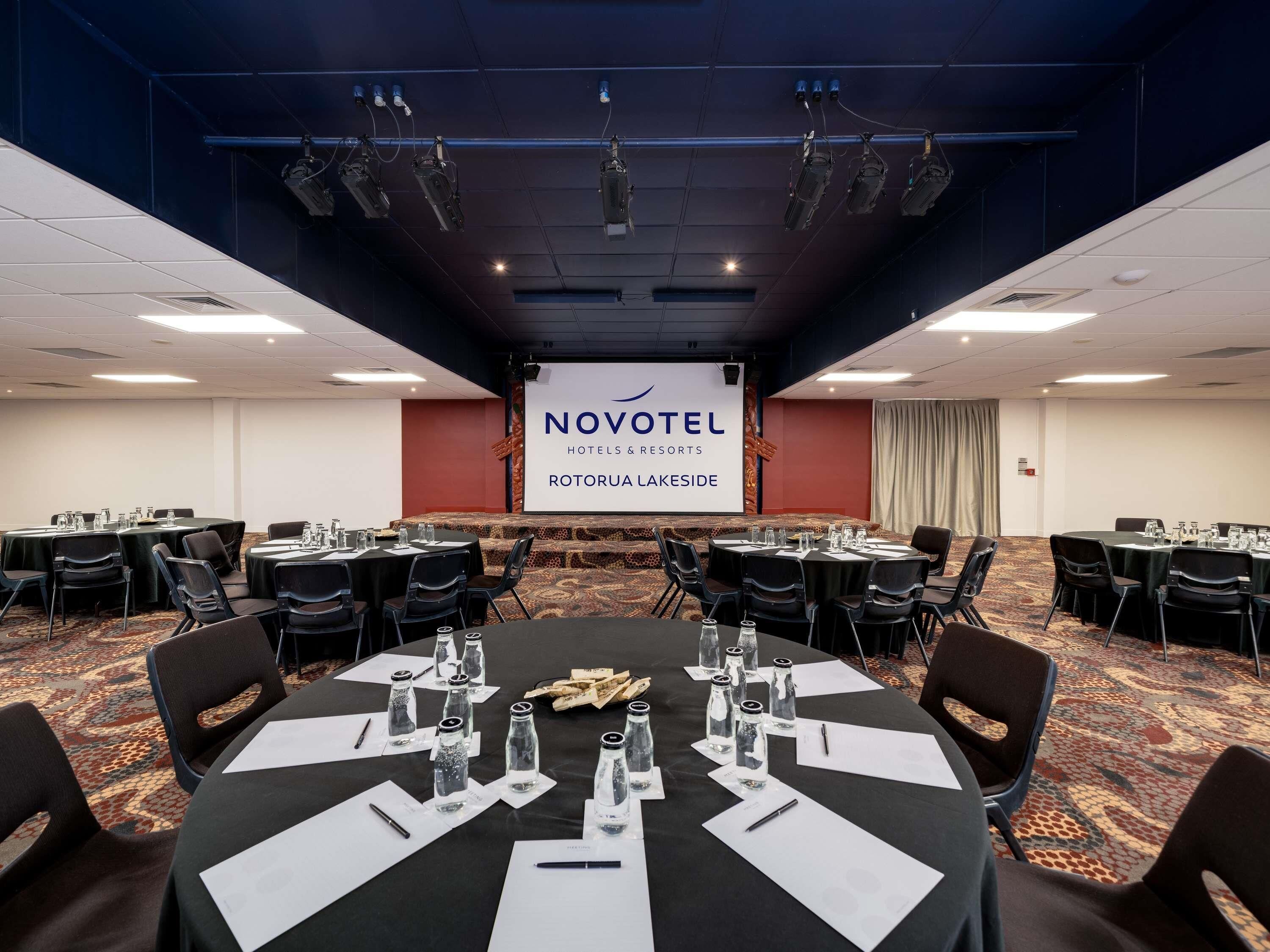 A conference room with tables, chairs, and a projector screen displaying 'NOVOTEL HOTELS & RESORTS ROTORUA LAKESIDE'.