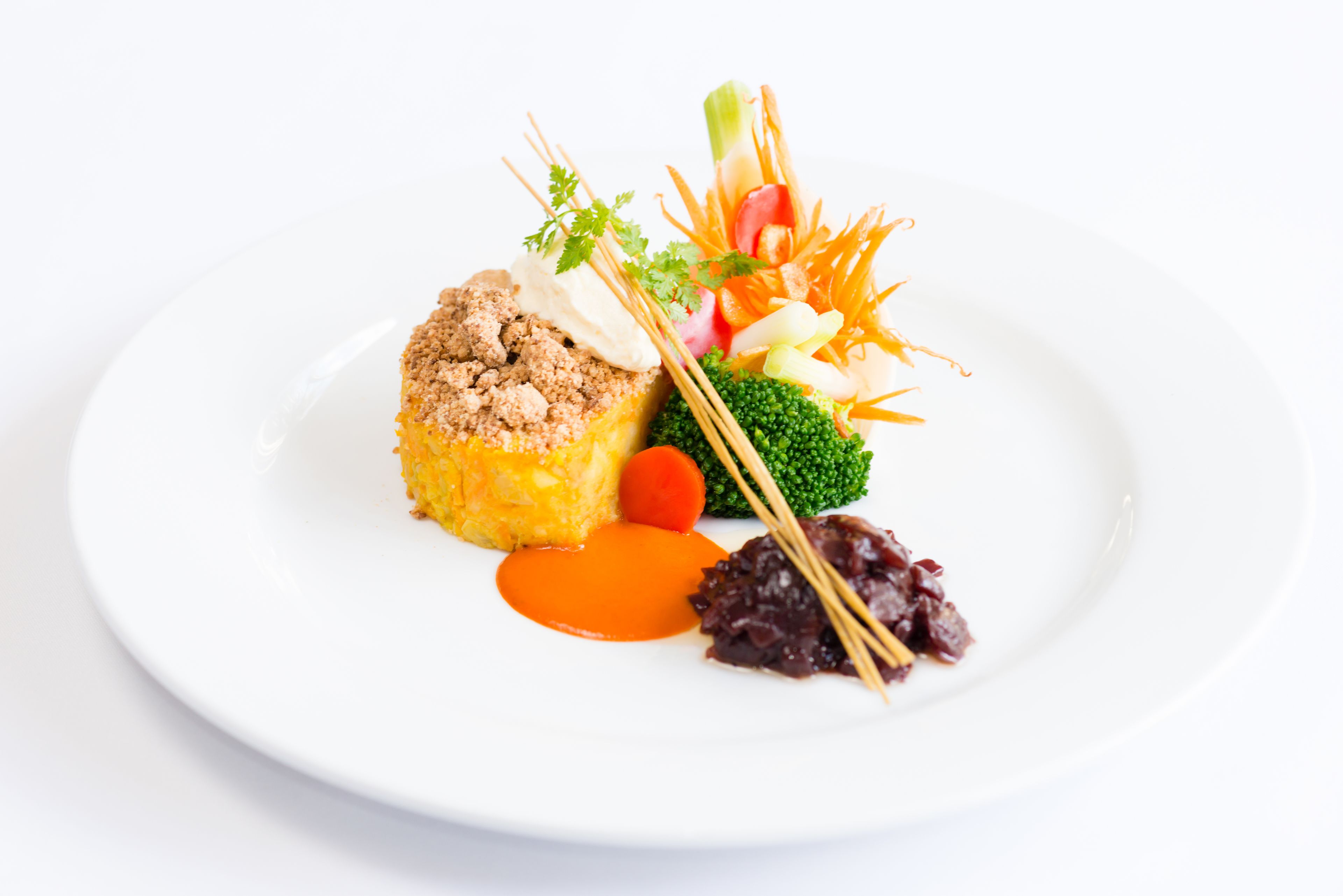 A white plate with a piece of food topped with breadcrumbs, a small carrot, a dollop of sauce, and a garnish of vegetables.