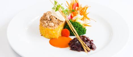 A white plate with a piece of food topped with breadcrumbs, a small carrot, a dollop of sauce, and a garnish of vegetables.
