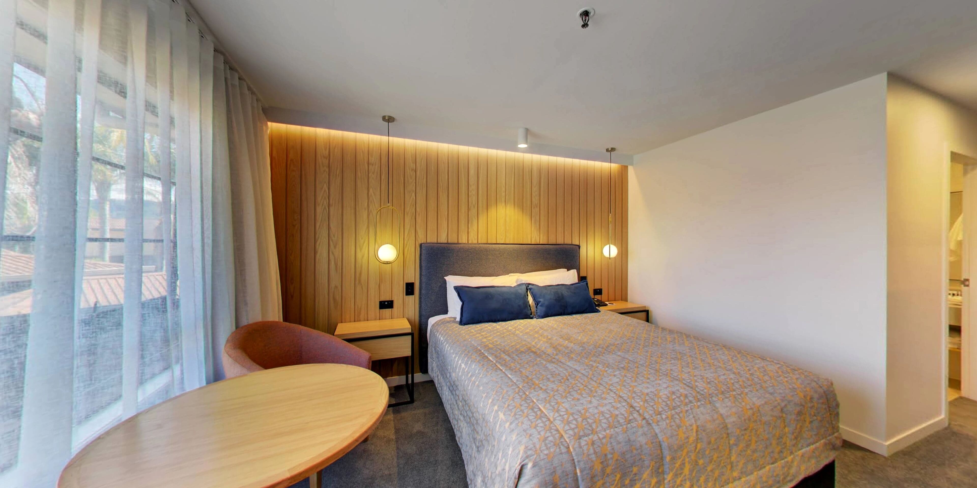 A modern hotel room with a large bed, a small table, a chair, and a view of the outdoors through the window.