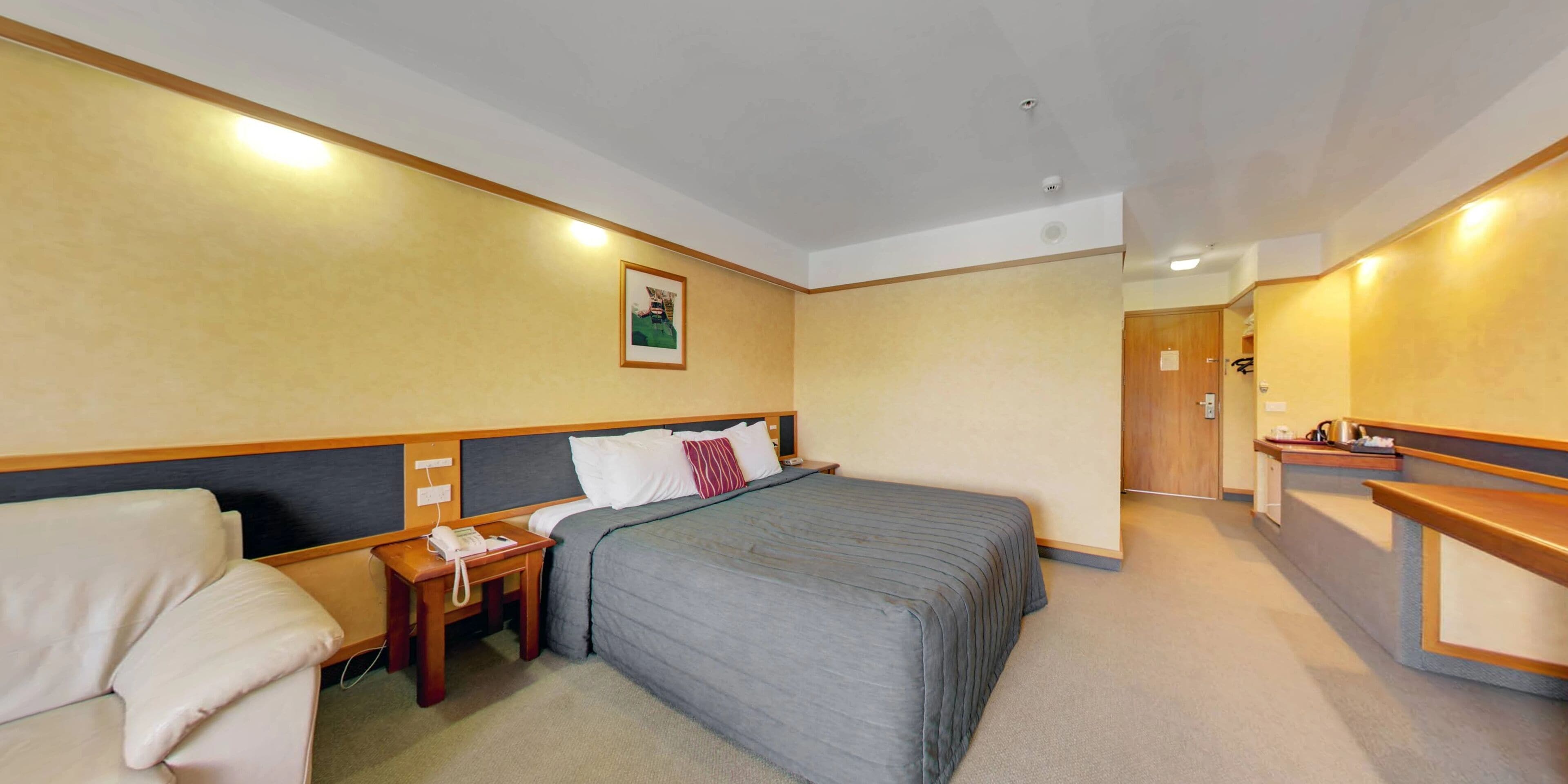 Quarto de hotel com cama, mesa de cabeceira, abajur e uma pequena escrivaninha com cadeira.