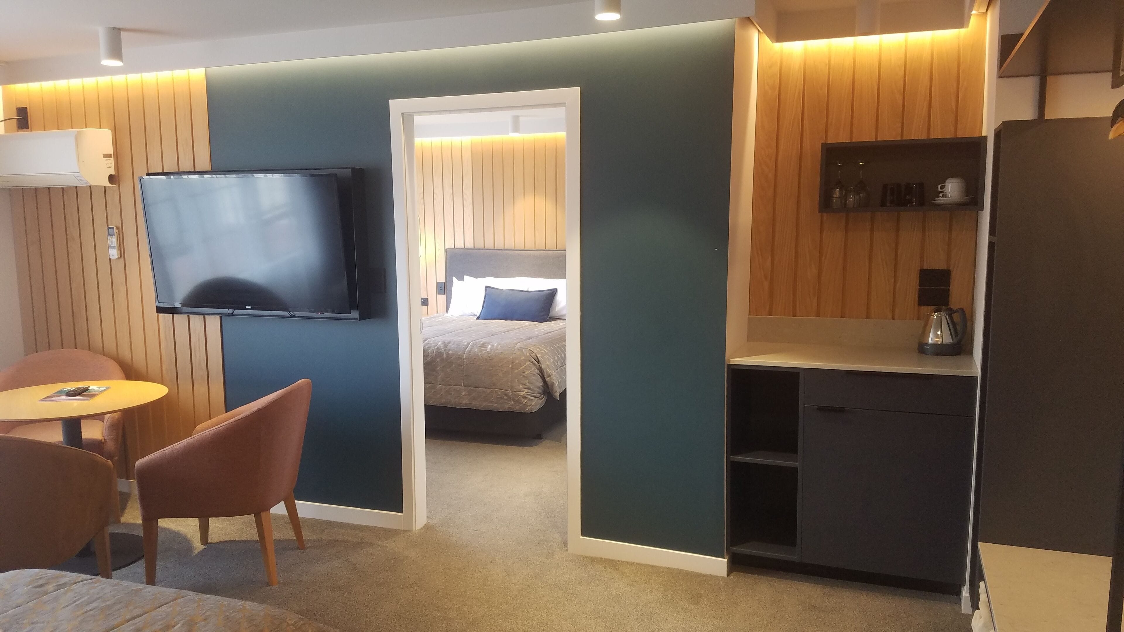 Una habitación de hotel con televisión, cama, kitchenette pequeña y zona de estar con mesa y sillas.
