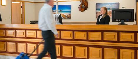 A hotel reception area with a wooden reception desk, a receptionist, and a man with a suitcase walking by.