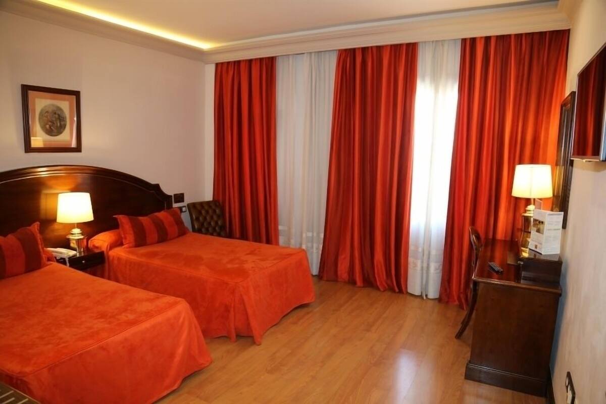 A hotel room with two beds, a wooden headboard, a desk with a lamp, and a large window with red curtains.