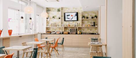 A modern restaurant with a long counter, multiple tables, and chairs. There is a TV mounted on the wall and shelves with various items.