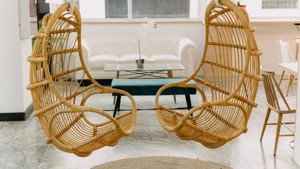 Una habitación con dos sillas colgantes de ratán, un sofá blanco, una mesa de centro de vidrio y una pequeña orquídea en maceta.