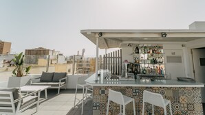 Un bar en la azotea con mostrador de bar de baldosas, taburetes blancos y una zona de estar con sofá y sillas.