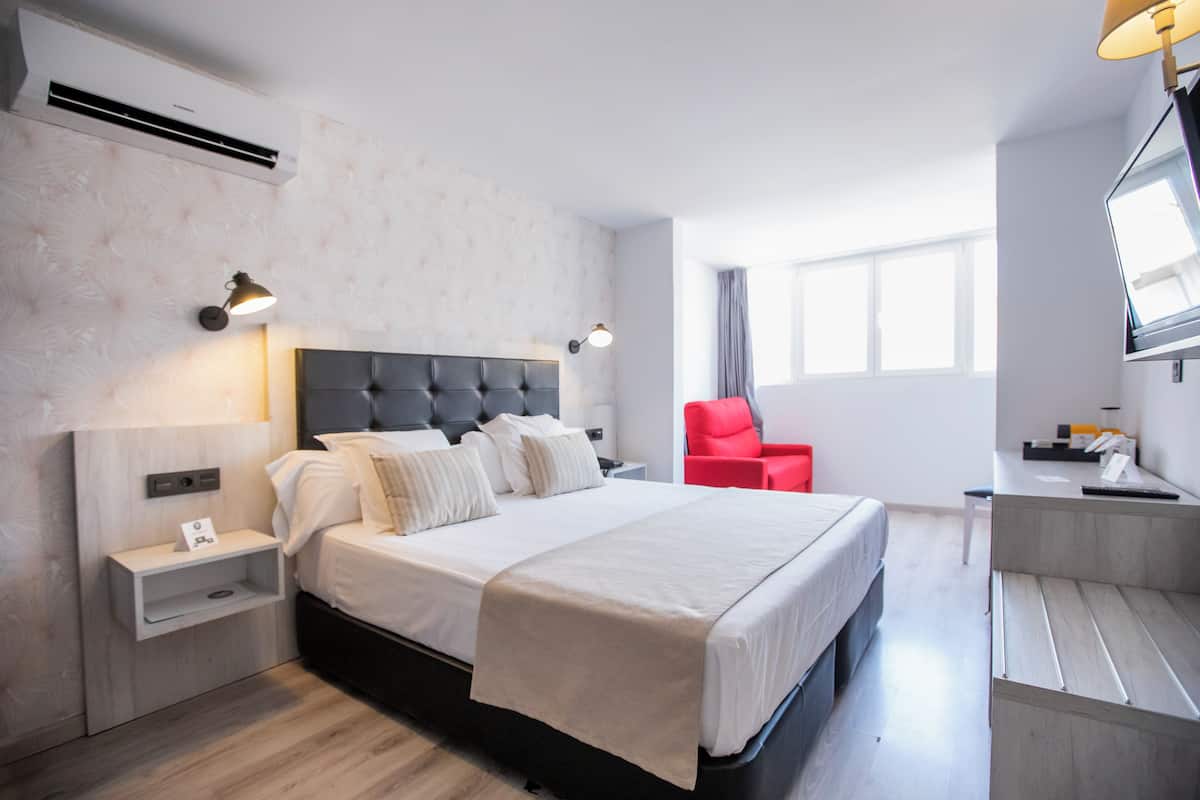 Quarto de hotel moderno com uma cama grande, mesas de cabeceira, uma escrivaninha e uma cadeira vermelha.